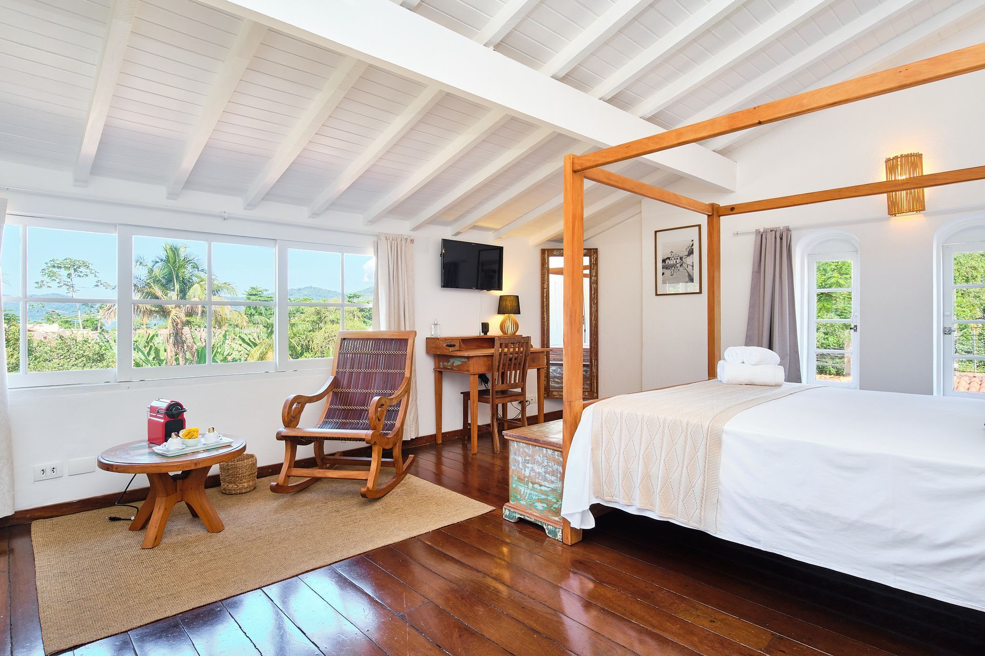Bright tropical bedroom with four-poster bed, lounge chair, and large windows overlooking greenery