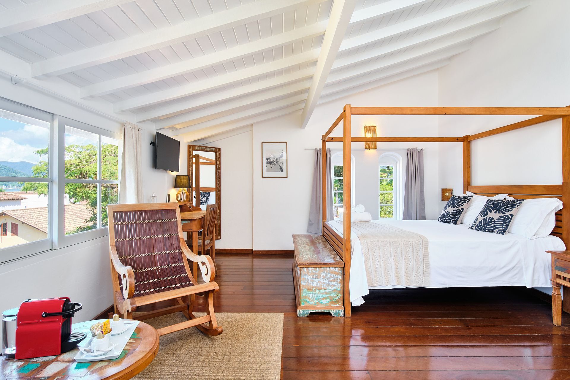 A bright bedroom with a wooden four-poster bed, rocking chair, and white sloped ceiling featuring exposed wooden beams.