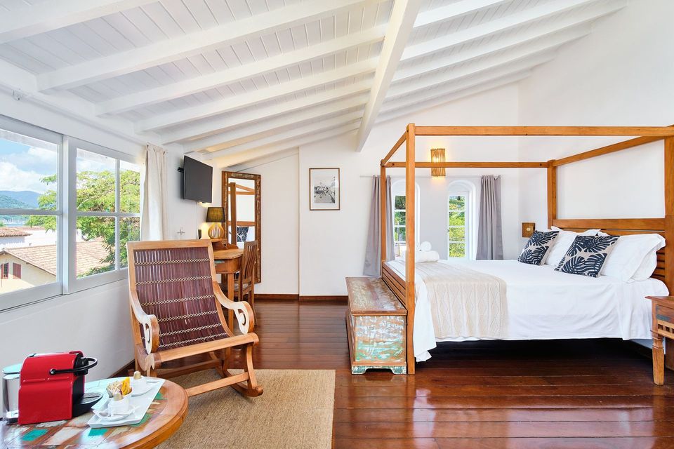 A bright bedroom with a wooden four-poster bed, rocking chair, and white sloped ceiling featuring exposed wooden beams.