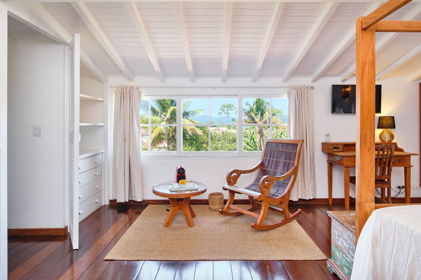 A bright bedroom featuring a wooden rocking chair, round coffee table, desk, and a view of tropical trees through windows.