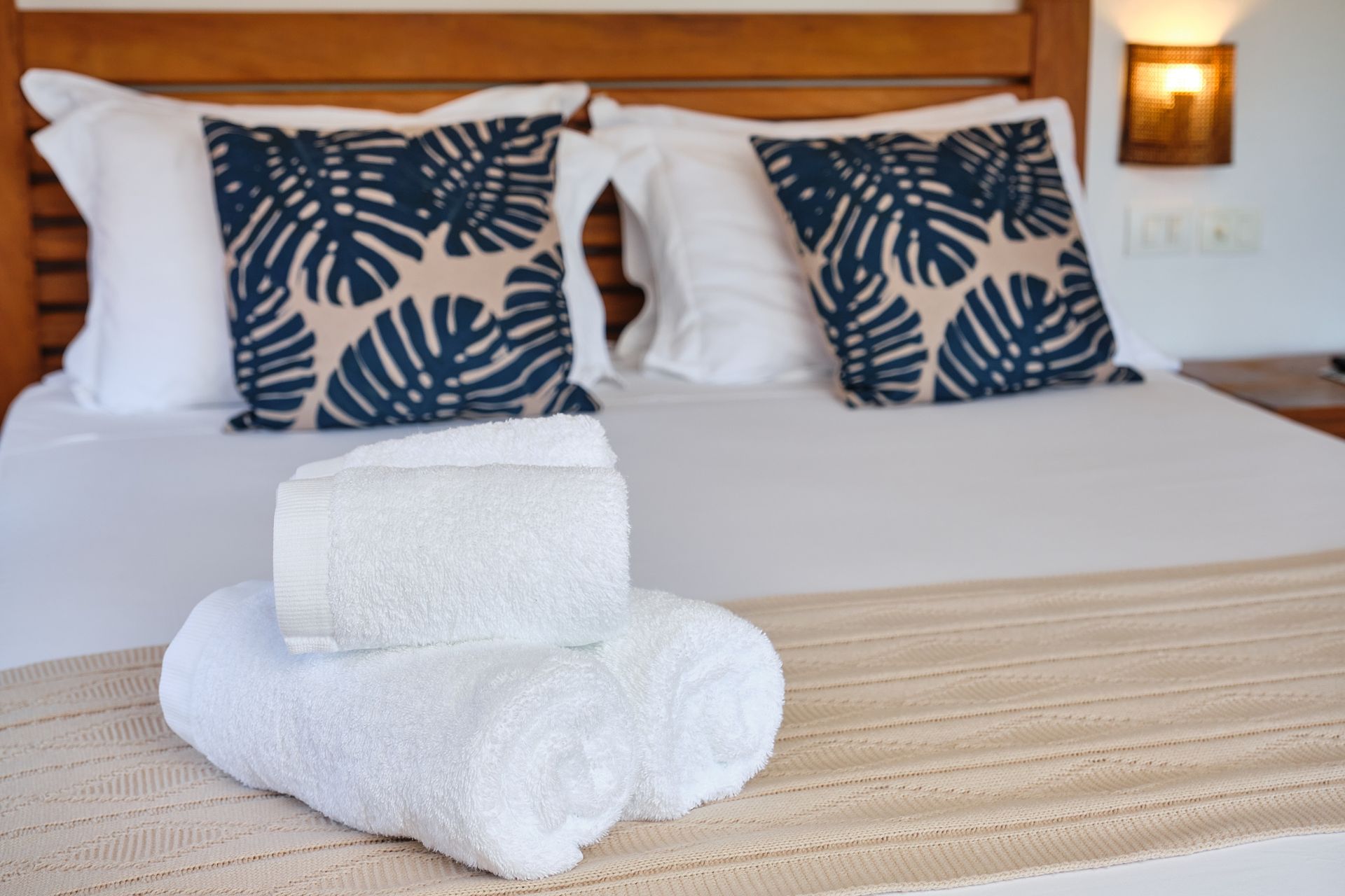 A neatly made bed with white linens, two palm-patterned throw pillows, and folded white towels on a textured runner.