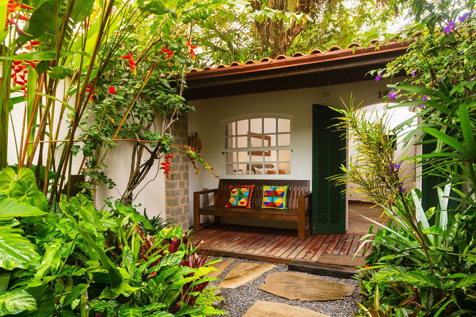 Garden cottage porch with wooden bench, colorful pillows, and lush tropical plants around it