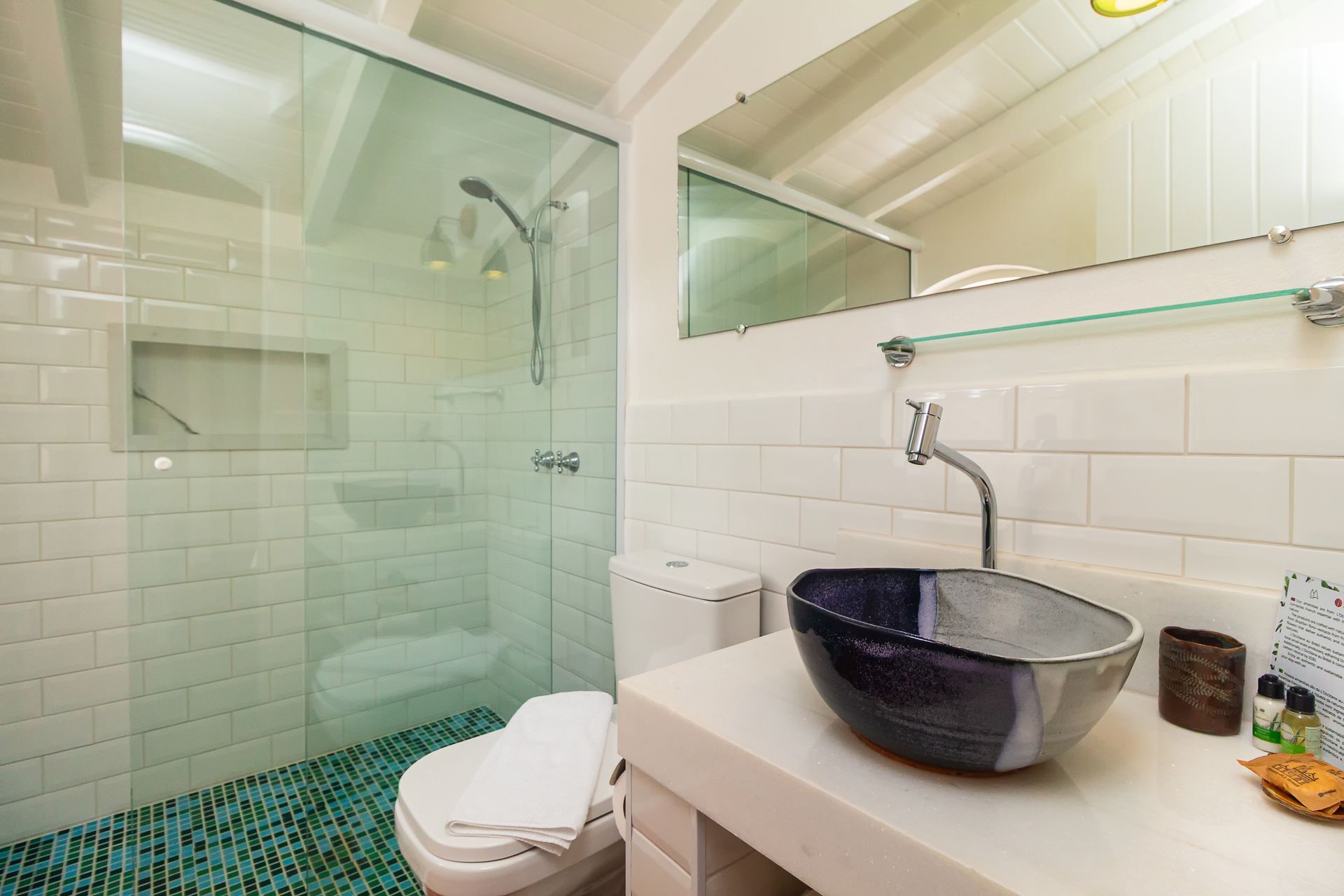 A modern bathroom with a glass-enclosed shower, subway-tiled walls, white toilet, and a blue and grey vessel sink.