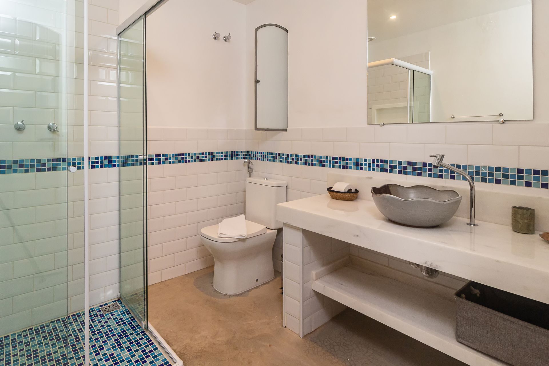 A white bathroom with a glass shower, a toilet, and a vanity featuring a grey bowl sink and blue mosaic tile accents.