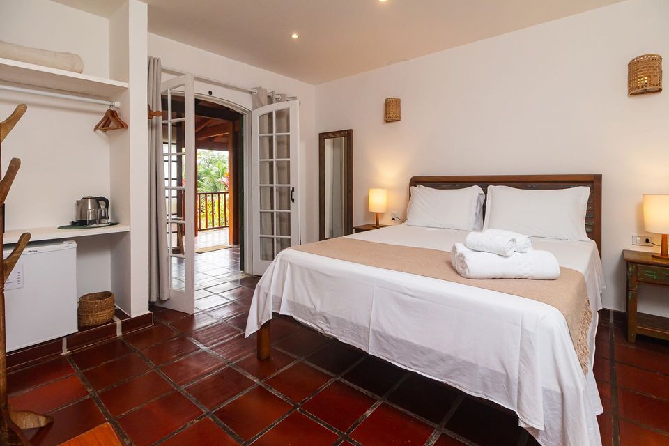 A hotel room featuring a queen bed with white linens, a terracotta tile floor, and French doors leading to a balcony.