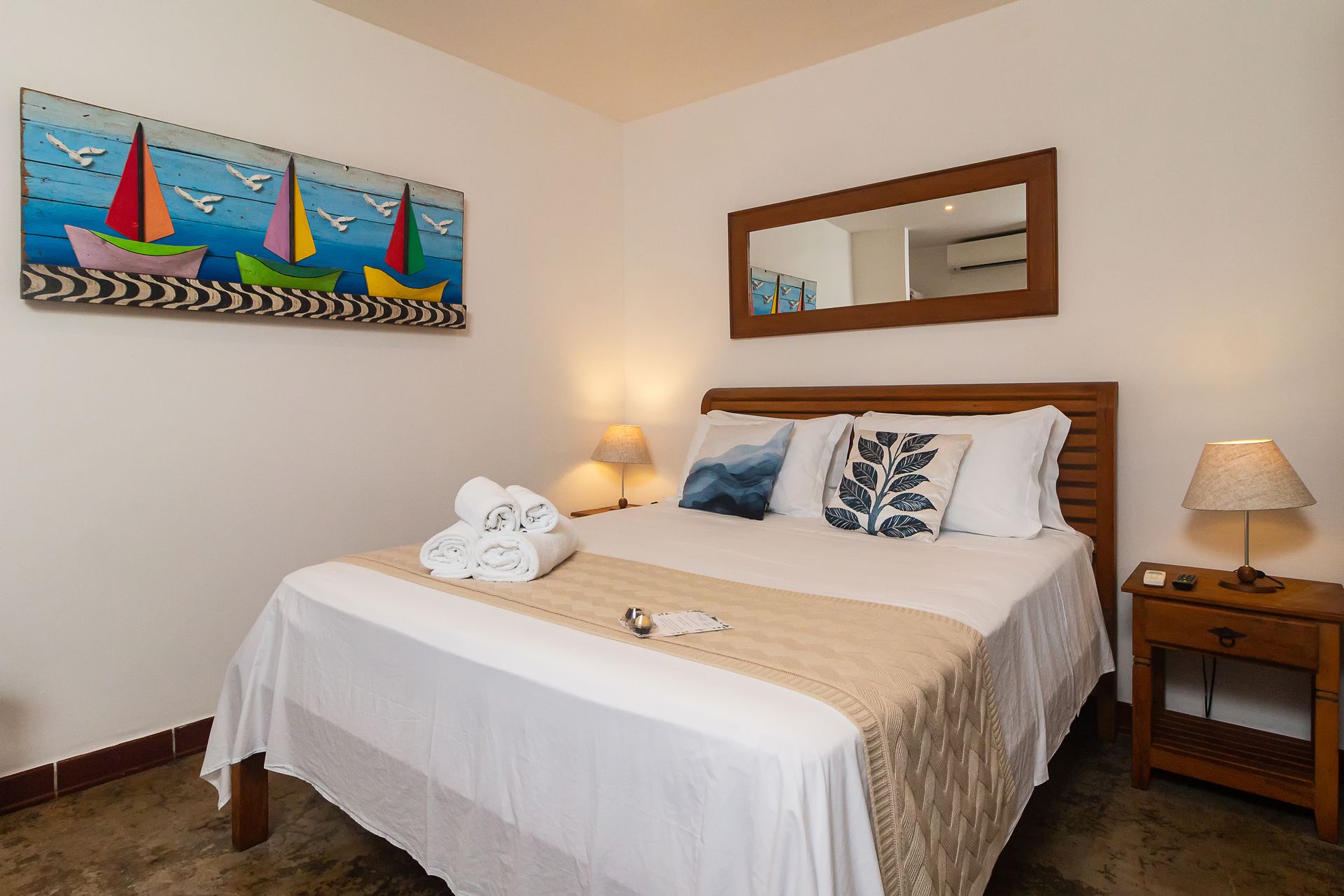 A hotel bedroom with a queen bed, nautical wall art, a wooden headboard, a mirror, and two bedside lamps.