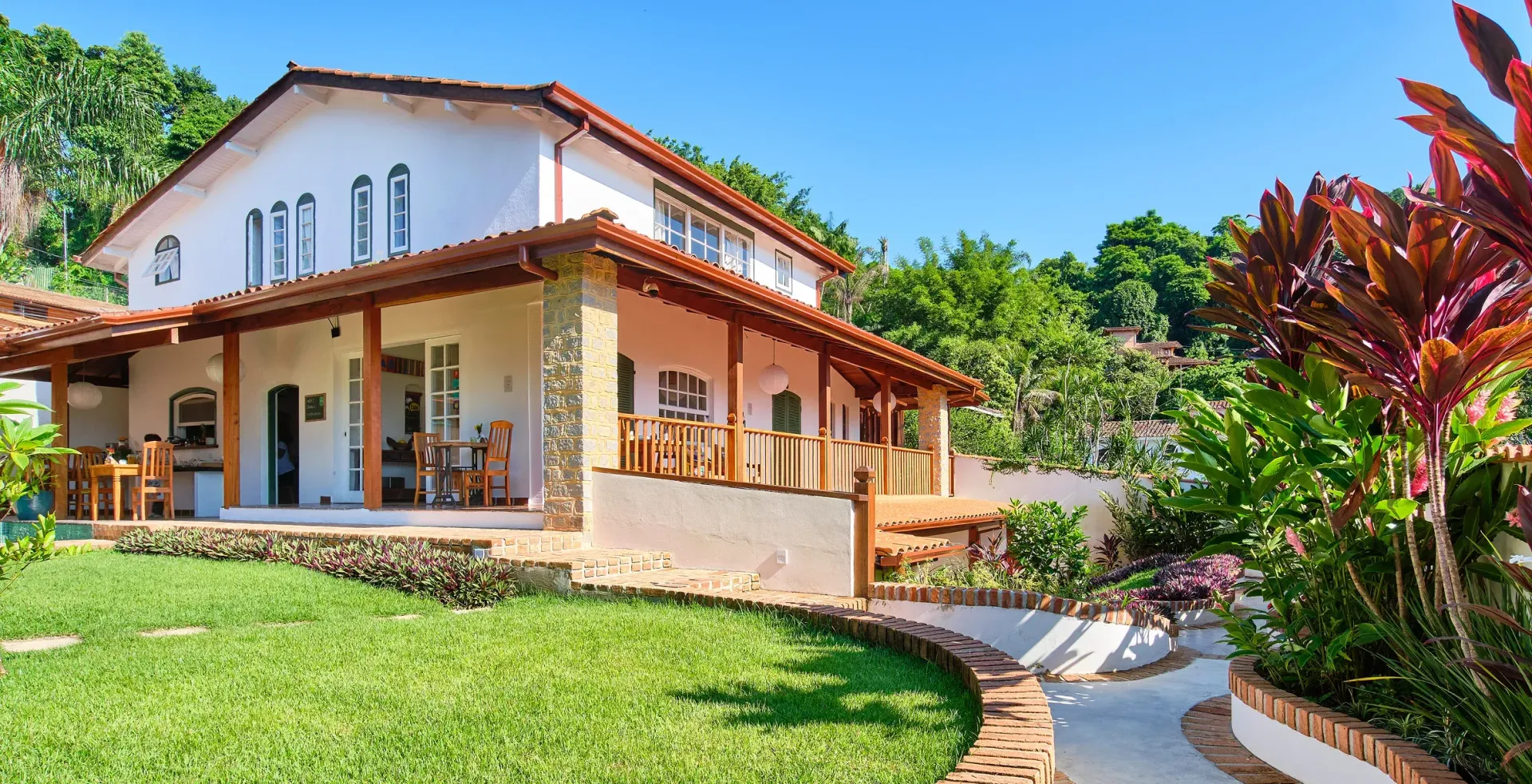 A two-story white house with a covered porch and terracotta tile roof, set in a lush garden with a curved walkway.