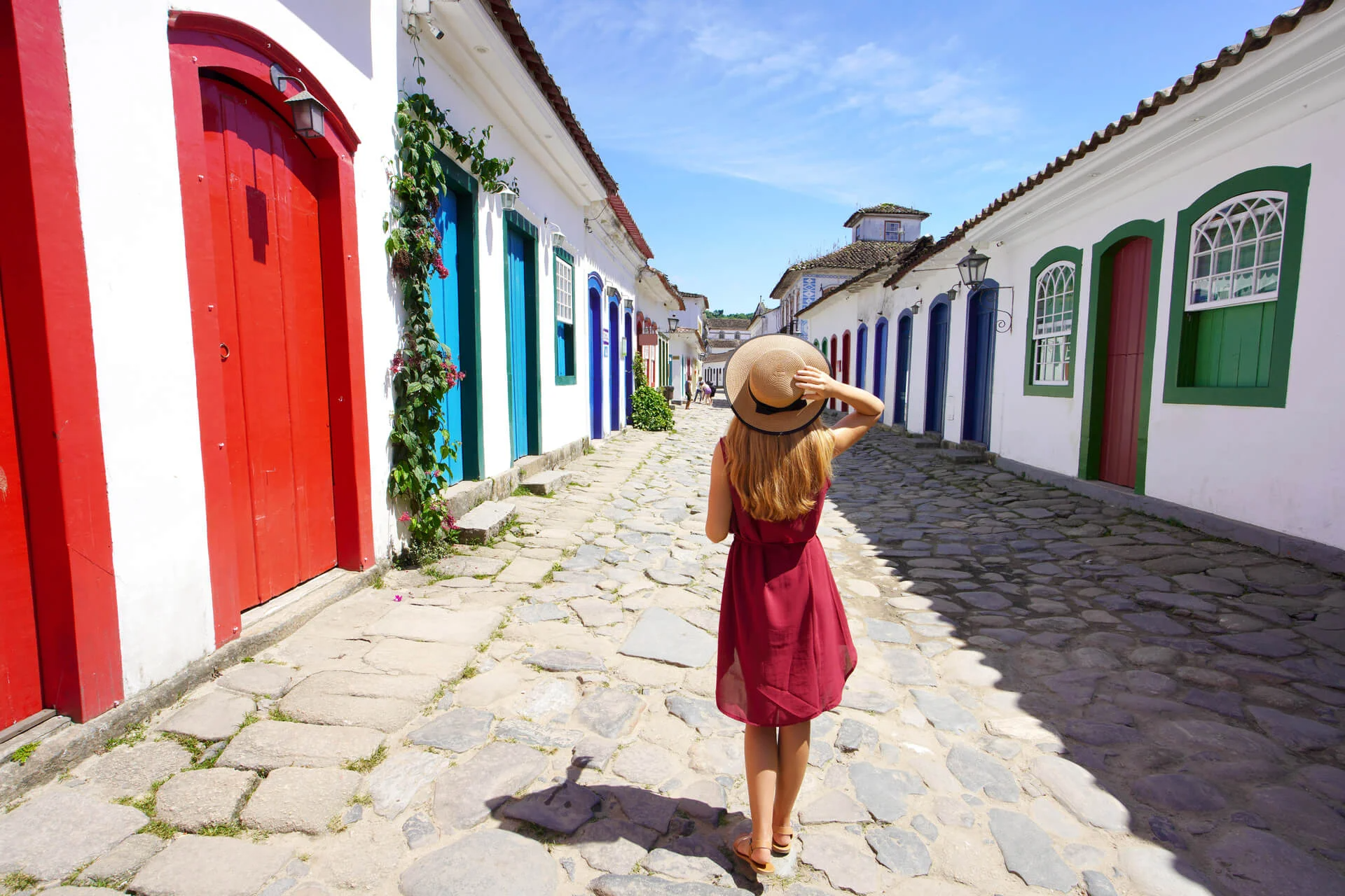 A person in a red dress and hat walks down a cobblestone street lined with colorful doors and white buildings.