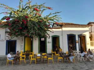 People sit at yellow chairs outside a white colonial-style building adorned with a large, flowering bougainvillea bush.