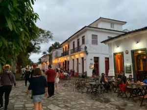 A cobblestone street in a colonial town at dusk, with people walking past white buildings and outdoor restaurant tables.