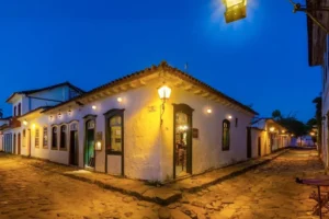 A historic white corner building illuminated by warm yellow lights at dusk, set along a cobblestone street.