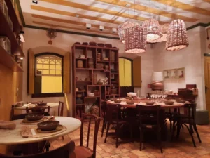 A warm, rustic dining area featuring round tables, wooden chairs, open shelves filled with pottery, and wicker lamps.
