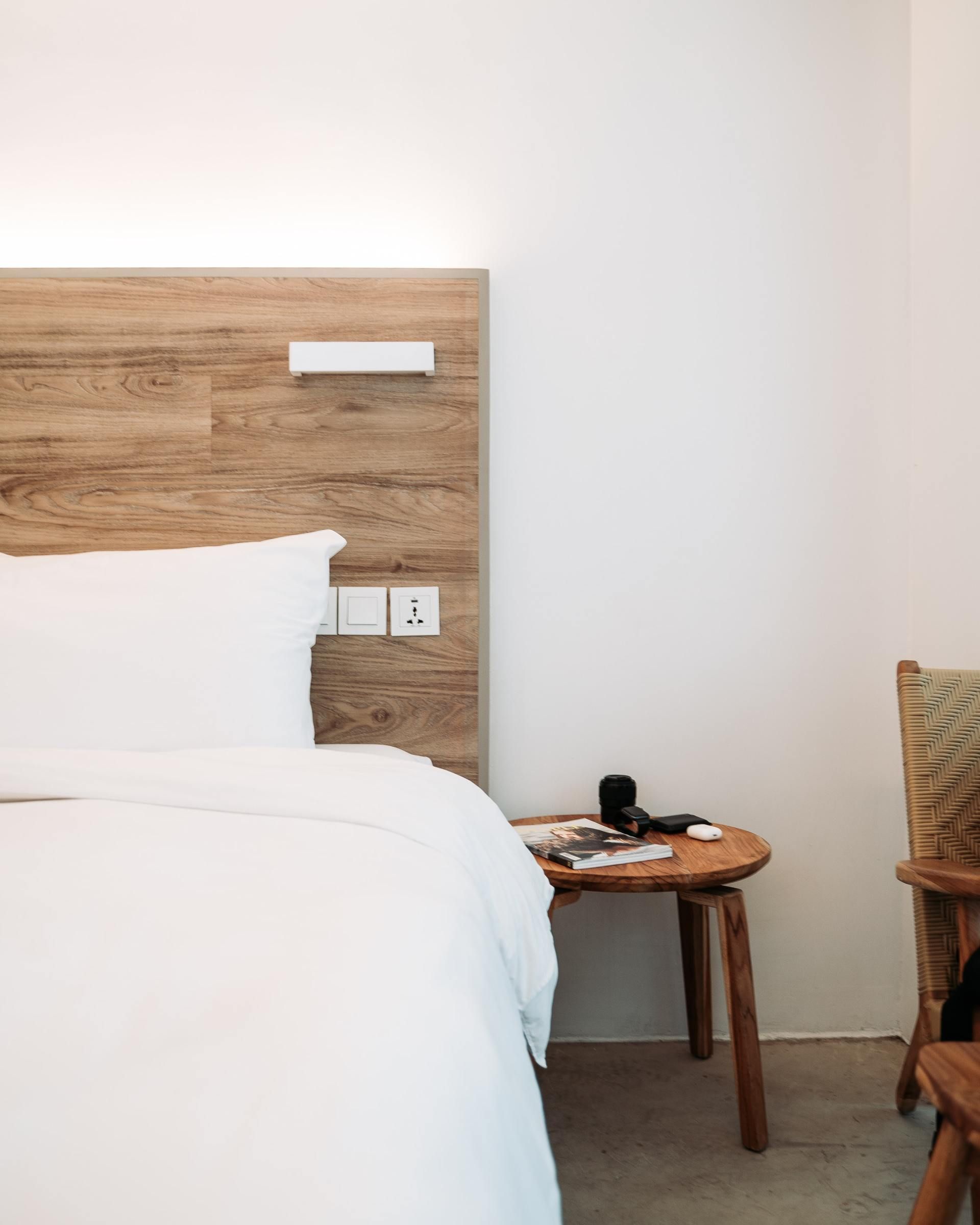 A minimalist bedroom featuring a white bed with a light wood headboard, a wooden side table, and a wooden chair nearby.