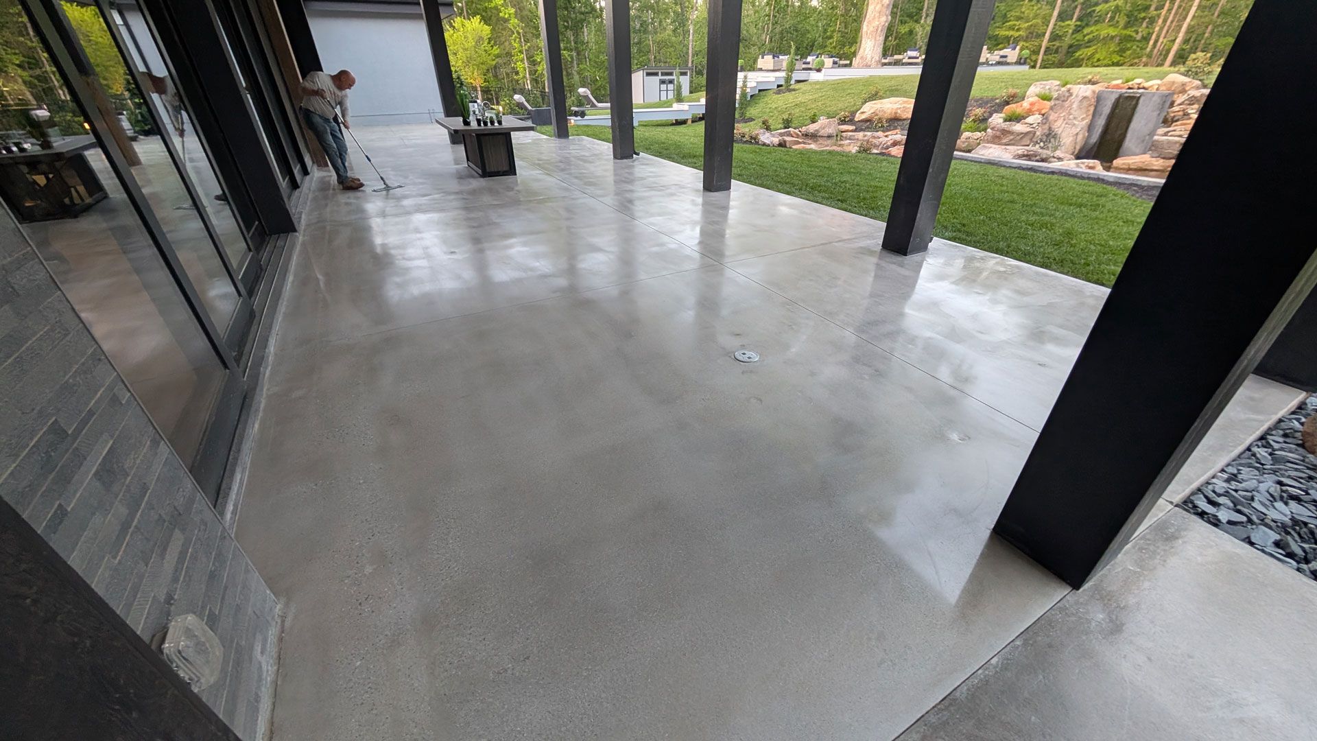 Empty covered patio with smooth concrete floor, dark support posts, and a view of a landscaped garden.