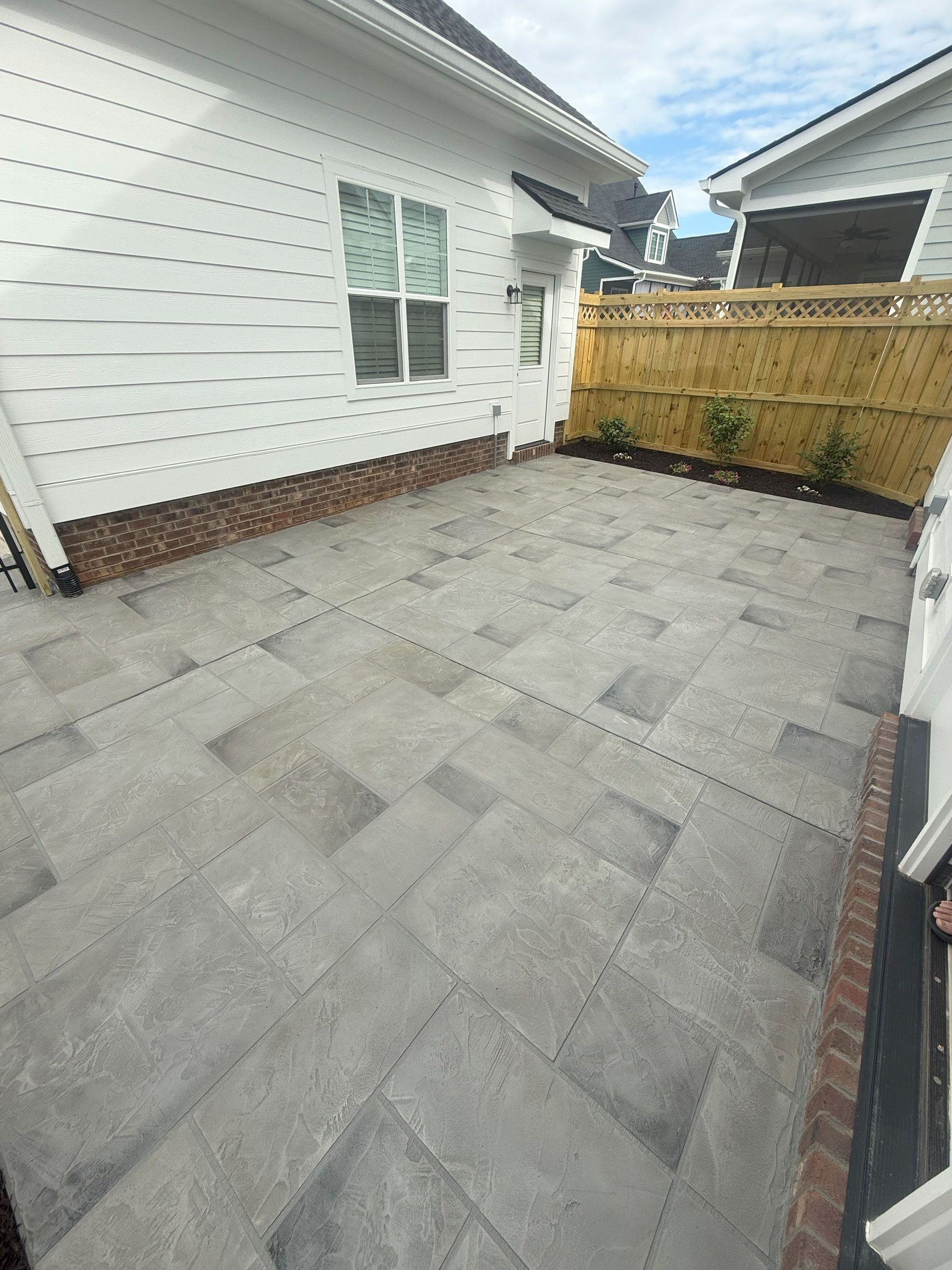 Decorative concrete patio with gray stamped overlay finish beside a white house and wooden fence in Greenville, SC.