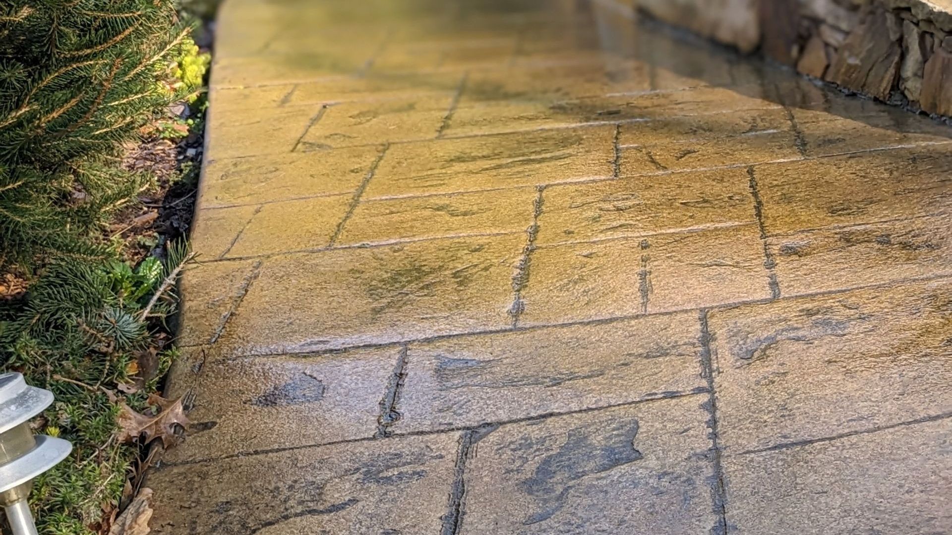 Stamped and sealed decorative concrete walkway with natural stone texture beside a garden bed and stacked stone wall.