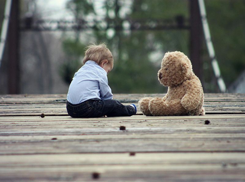 Small toddler boy with large Teddy Bear sitting on a deck.