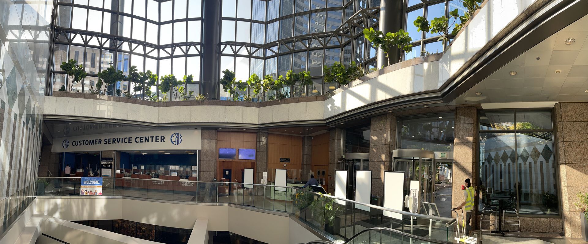Modern indoor mall atrium with glass roof, storefronts, and a central escalator under greenery
