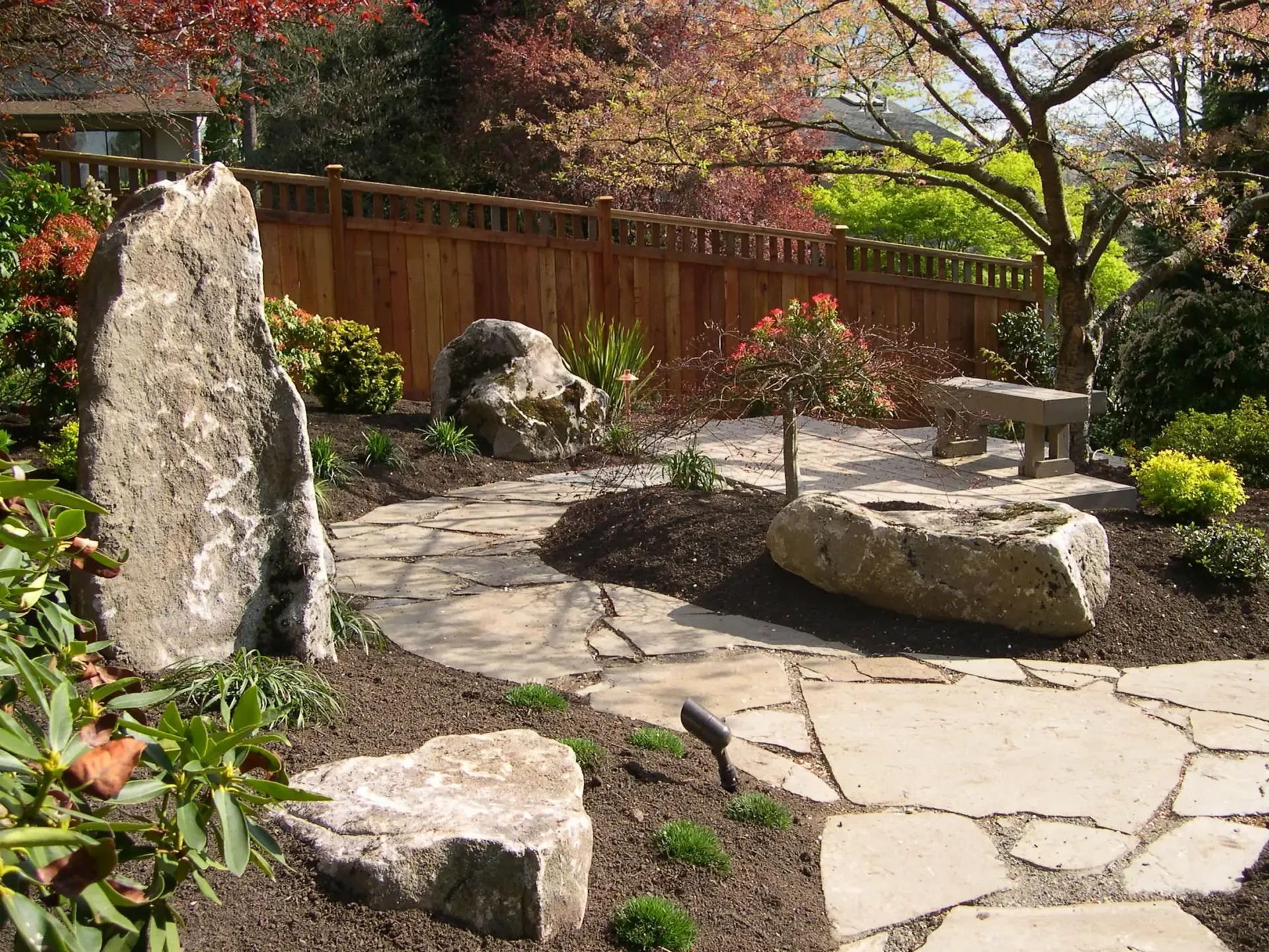 Stone garden path with large rocks, wooden fence, and blooming shrubs in sunlight