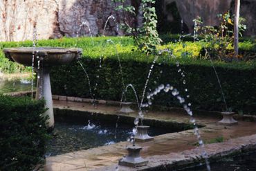 Garden fountain spraying water beside a stone birdbath and green hedges