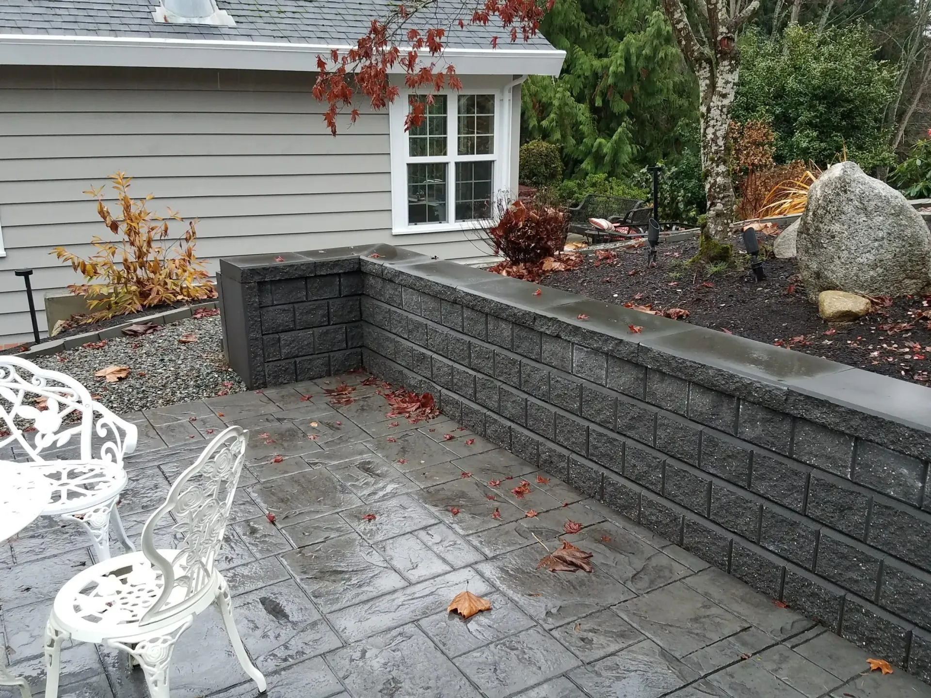Stone patio beside gray house with low retaining wall, white patio chair, and fallen leaves.