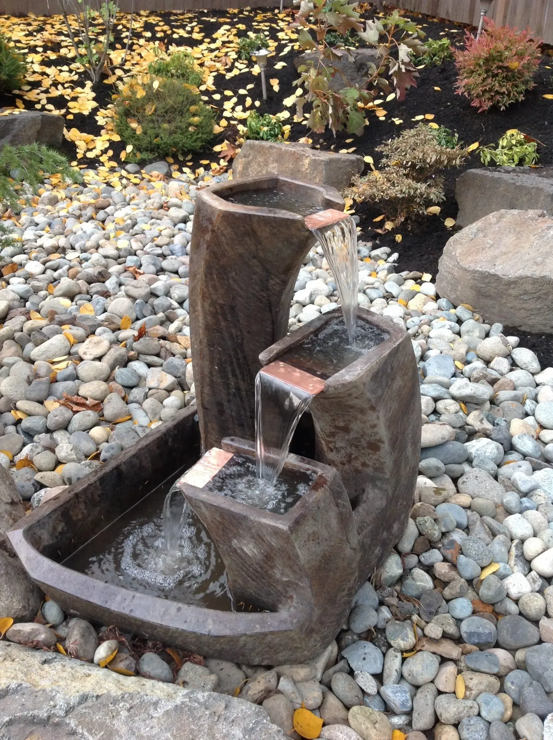 Small stone garden fountain with water flowing into a basin, set among pebbles and plants.