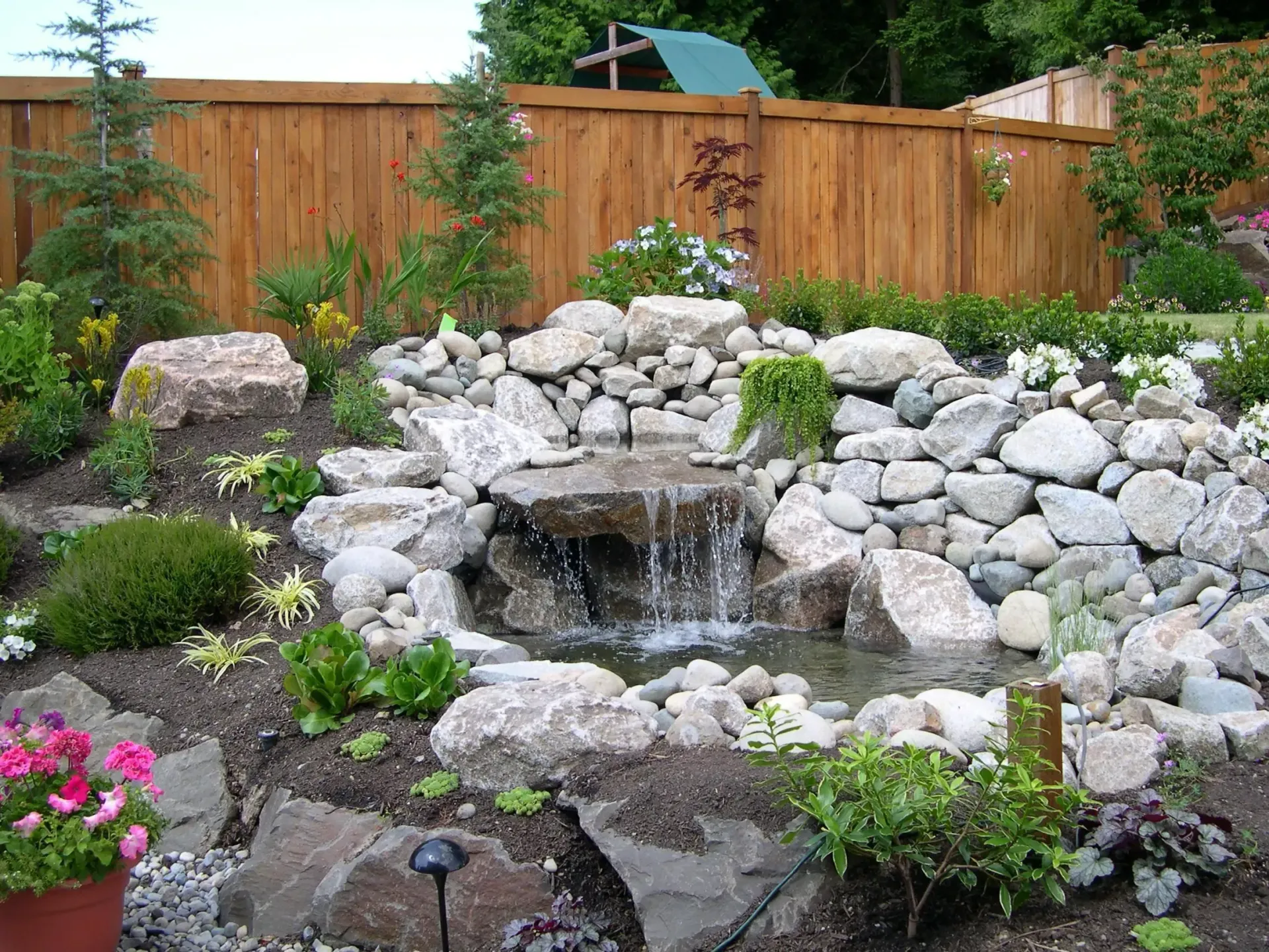 Backyard stone waterfall garden with rocks, flowers, and a wooden fence