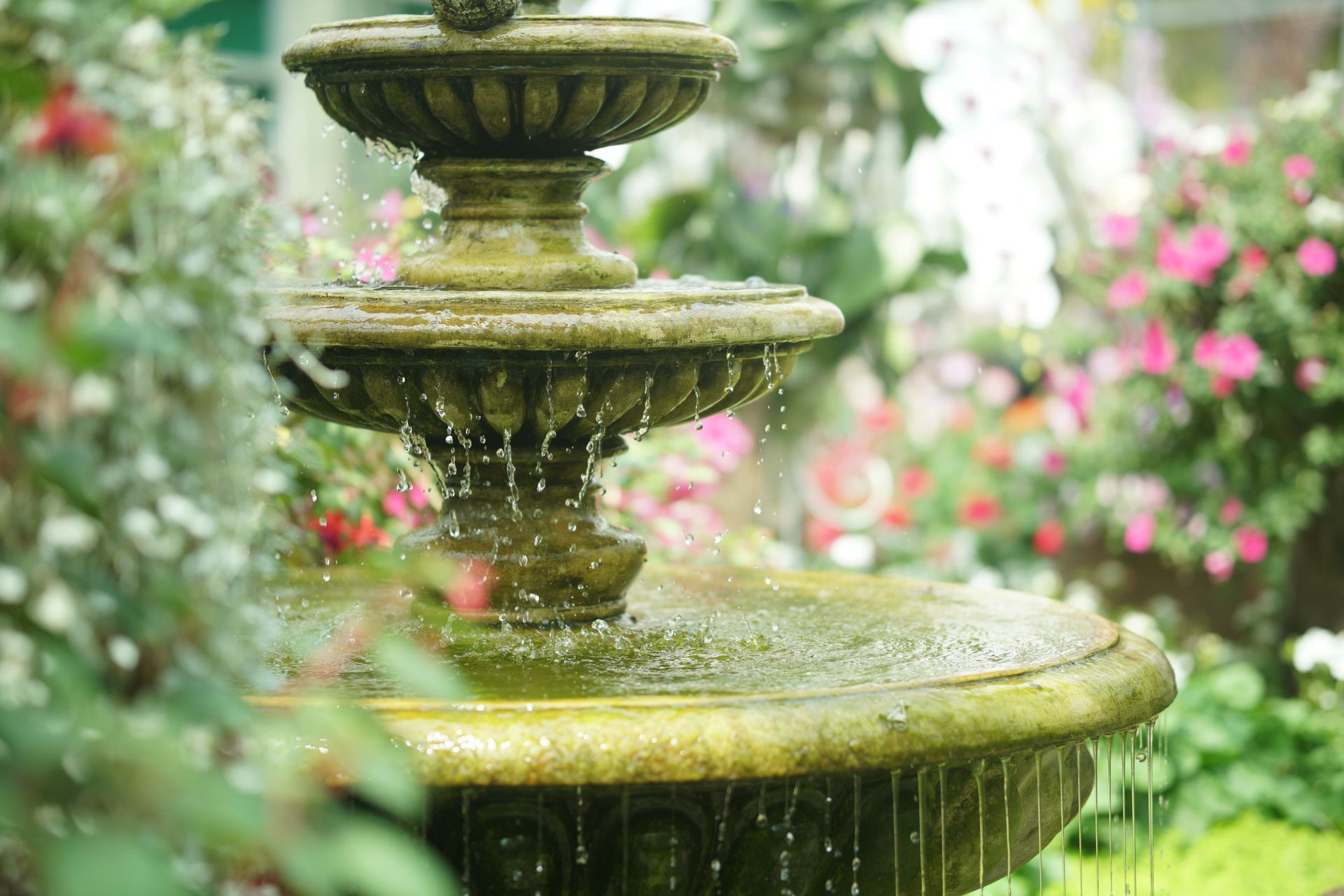 Tiered stone fountain with flowing water in a lush garden, surrounded by pink flowers and greenery