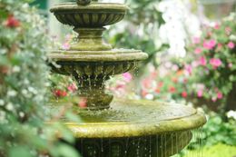 Tiered stone fountain with flowing water in a lush garden, surrounded by pink flowers and greenery
