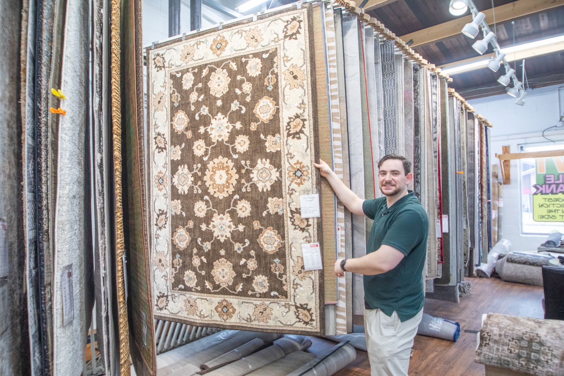 Man holding a large brown and cream patterned rug in a rug store. Rows of rugs are visible.