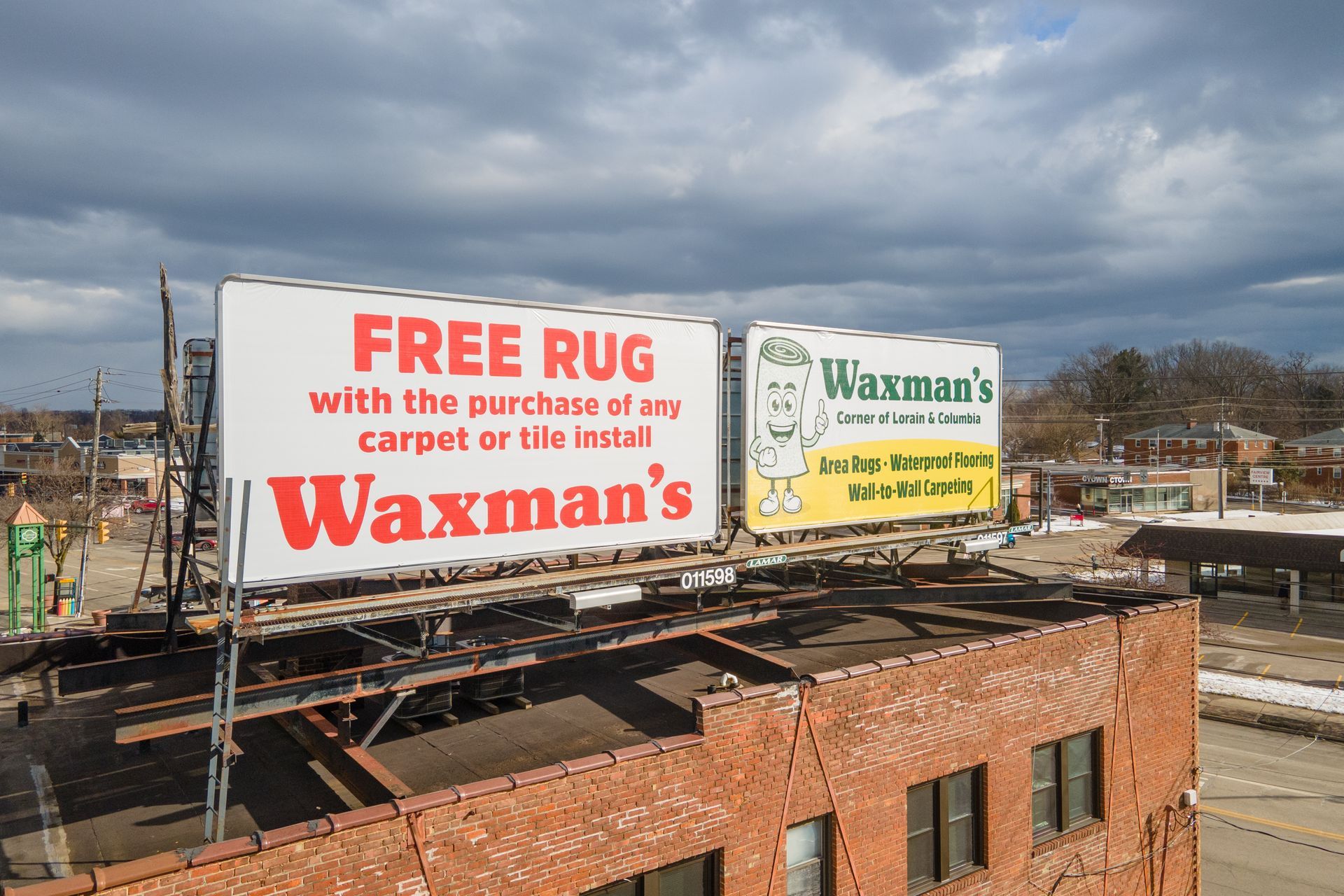 Billboard for Waxman's offers a free rug with purchase. On a rooftop, text and logo are visible. Cloudy sky.
