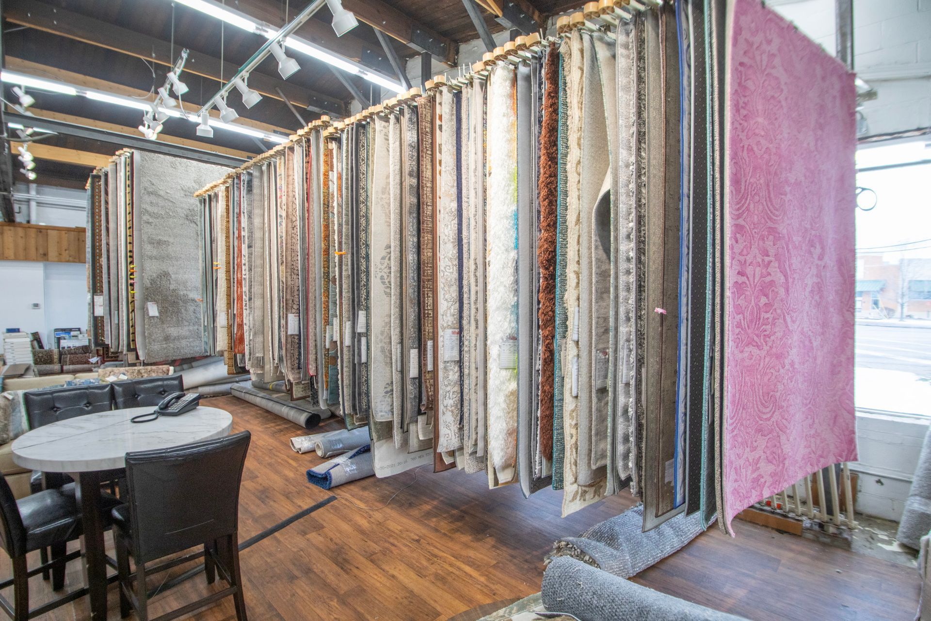 Carpet samples displayed in a showroom. Tables and chairs are in the foreground, and a window is visible.