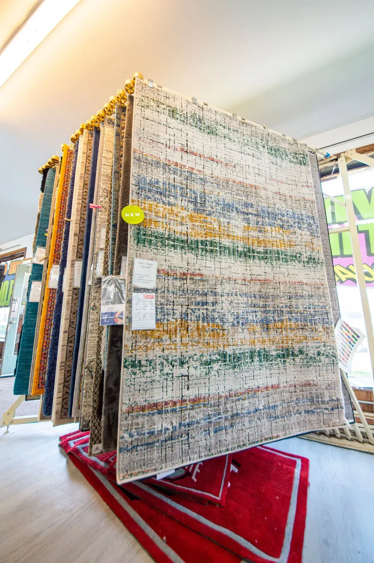 Rugs displayed in a store, leaning on wooden racks. The rugs are various colors and patterns, a red rug is on the floor.