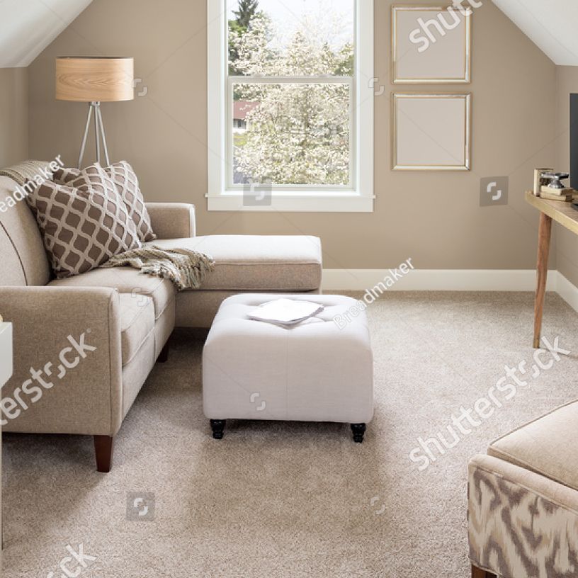 Beige living room with sectional sofa, ottoman, window, lamp, and neutral carpet.