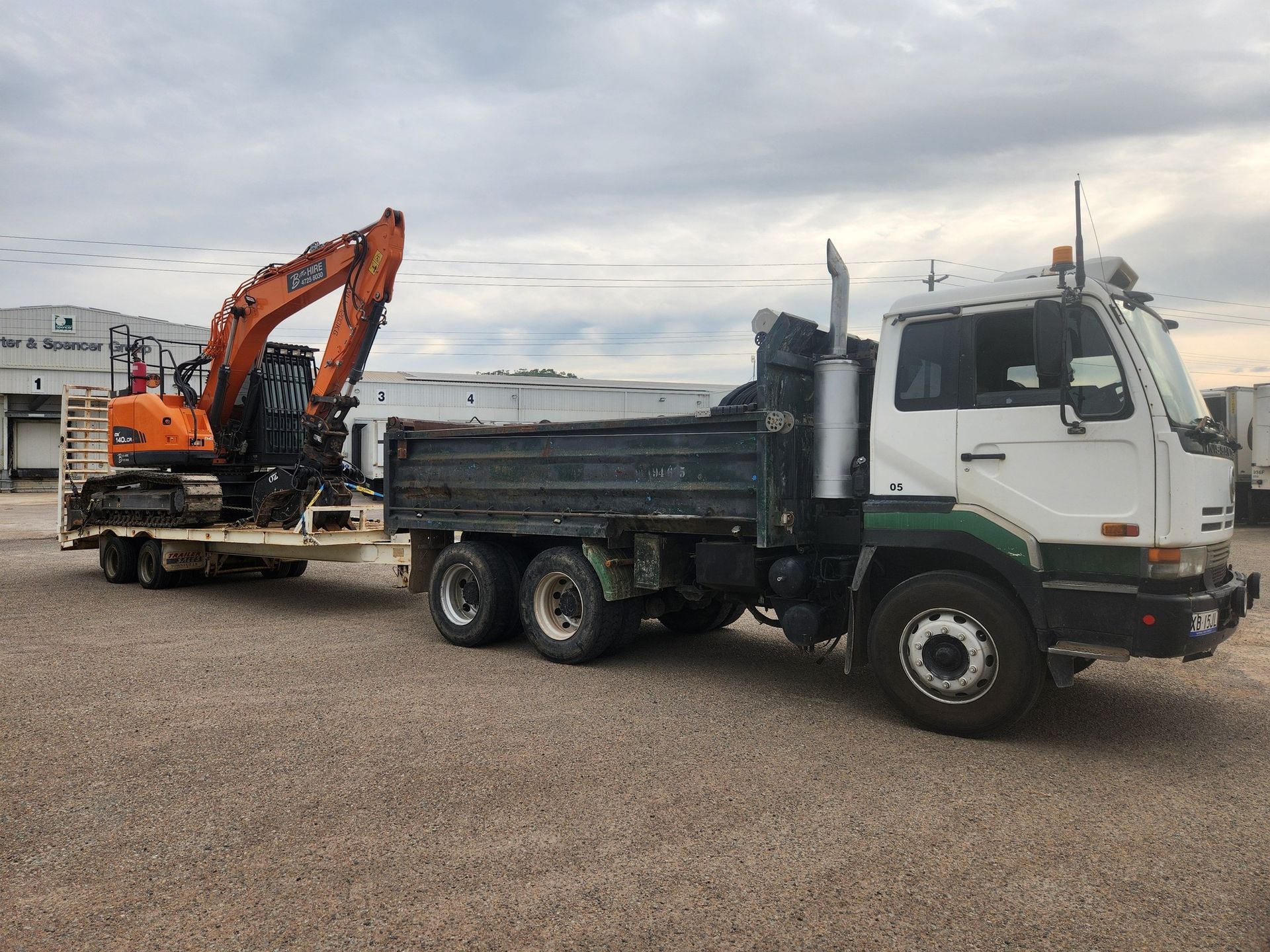 Truck With Excavator on Trailer, Parked on Gravel Lot Under Cloudy Sky — AGM HIRE NQ in Condon, QLD