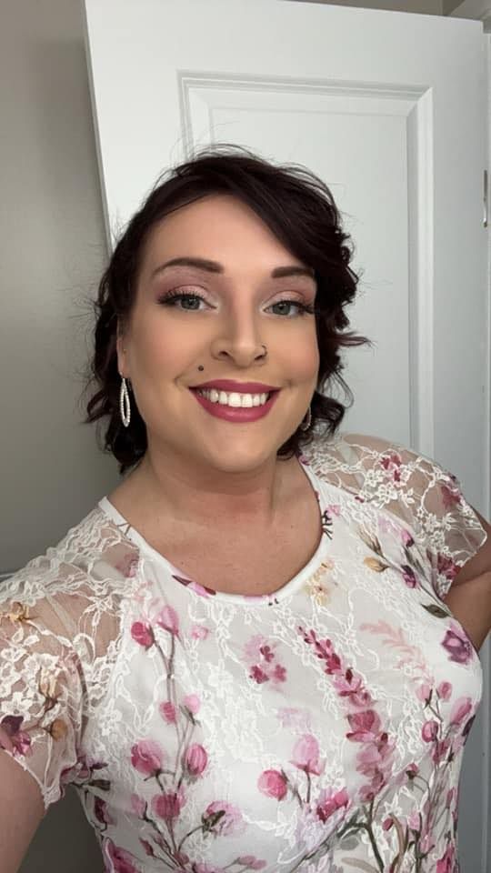 Smiling person with short dark hair in a white floral top standing indoors by a white door