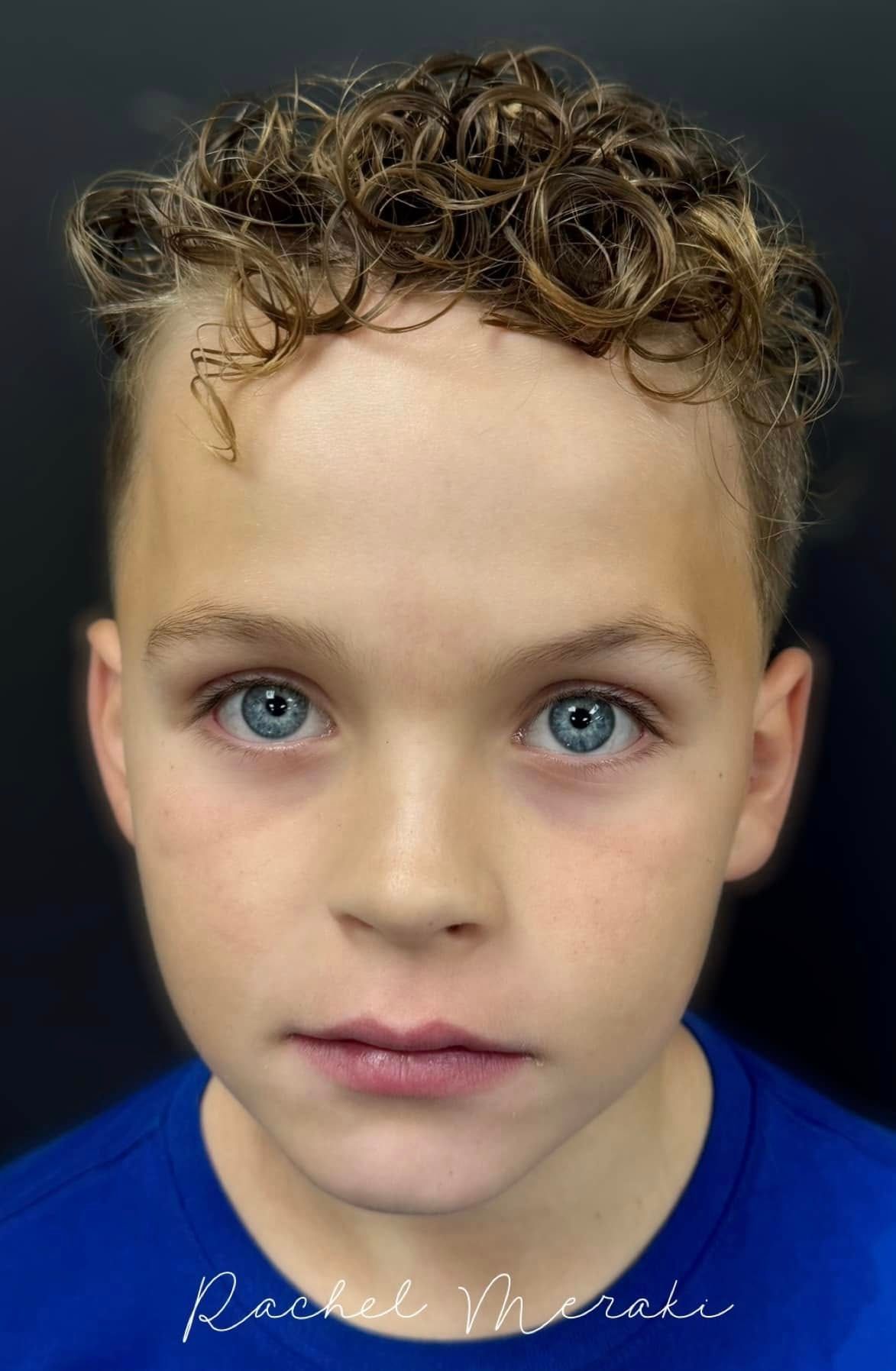Close-up portrait of a young person with curly hair and blue eyes against a dark background.