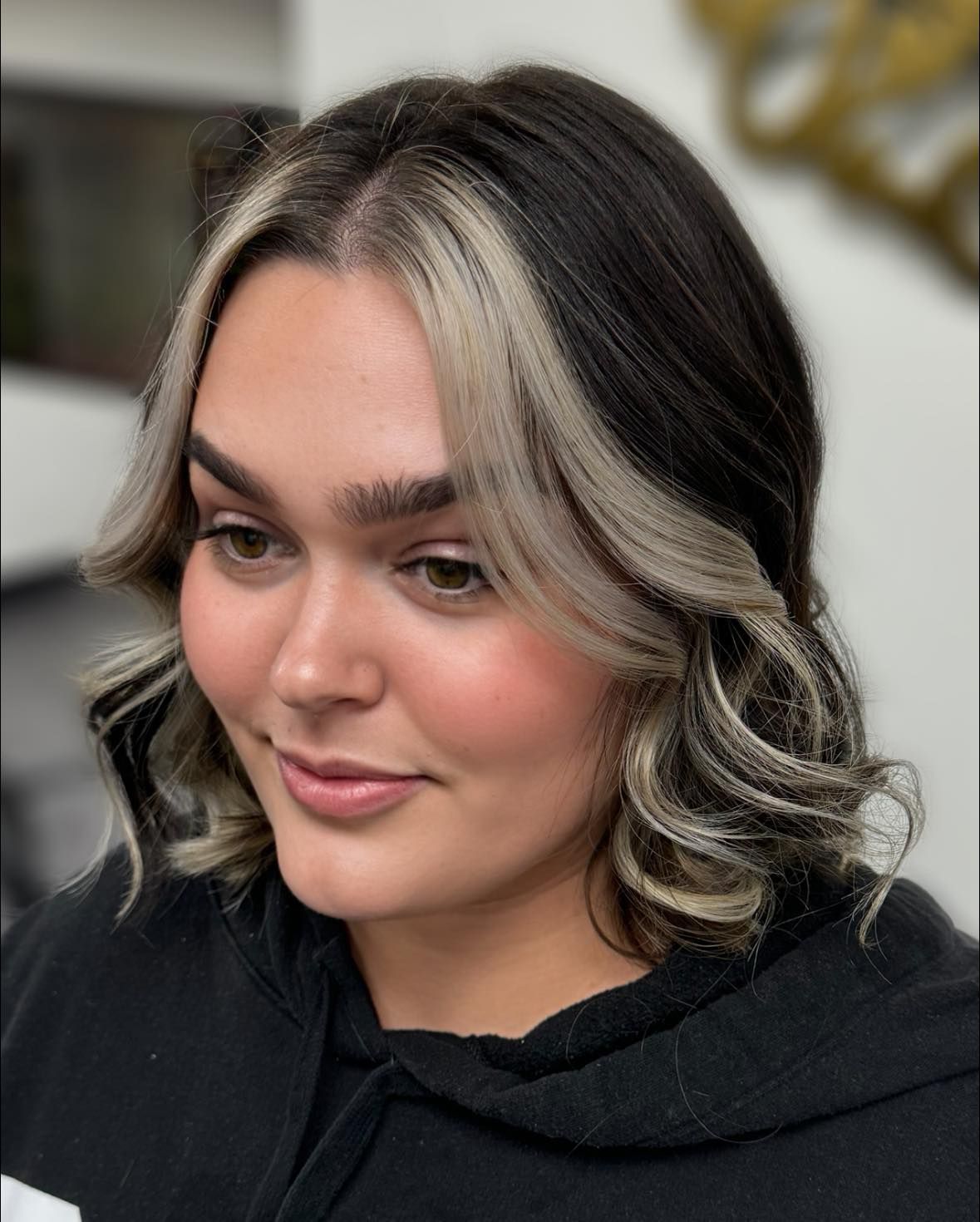 Portrait of a person with shoulder-length dark hair and blonde highlights, wearing a black top, softly smiling