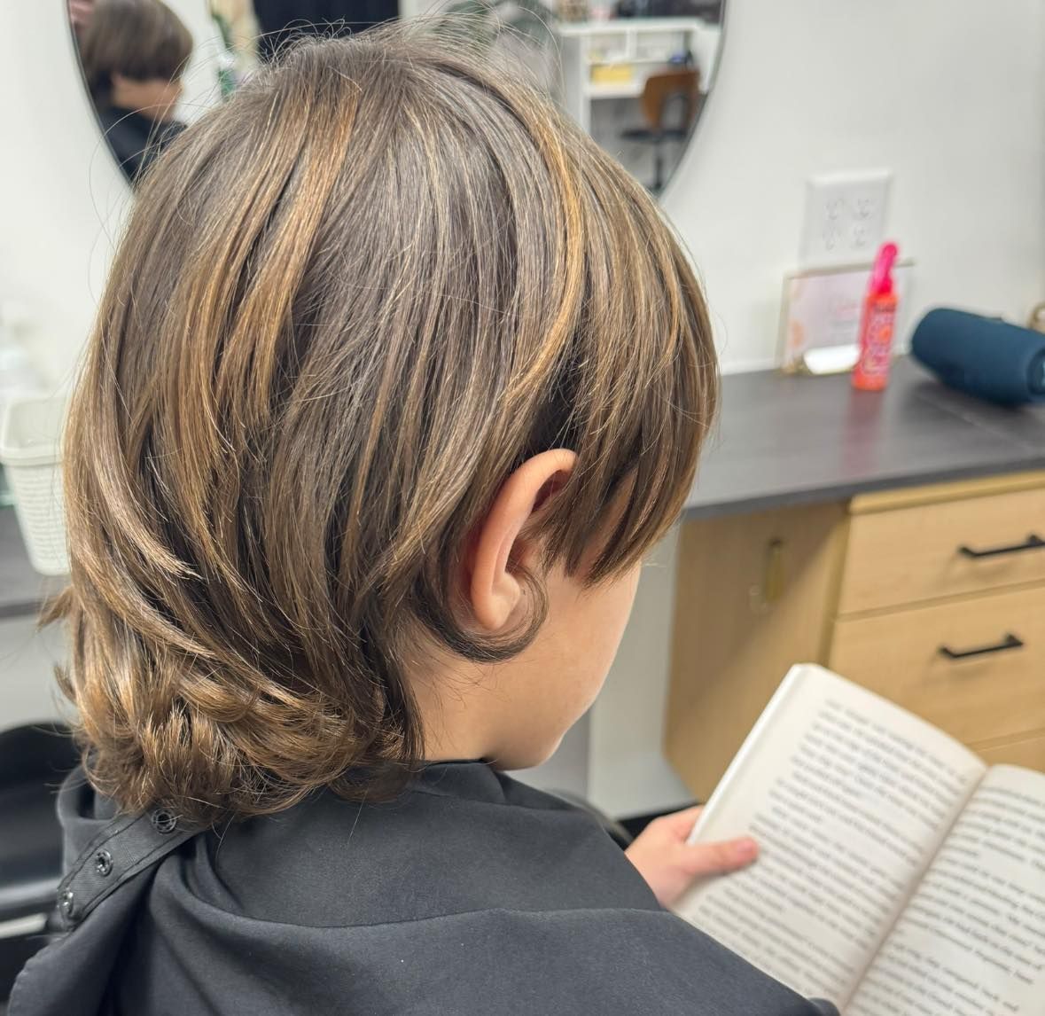 Child reading a book in a salon chair, seen from behind, with a mirror and countertop in the background