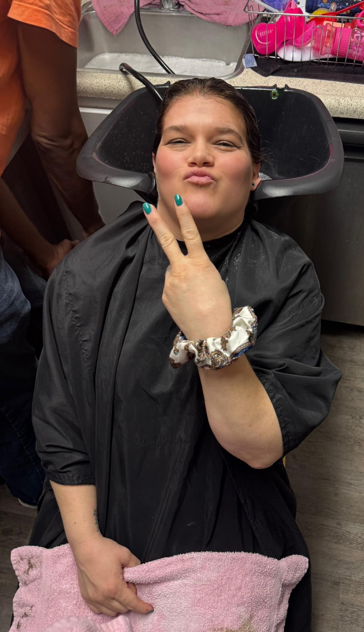 Person in a black outfit sitting and making a peace sign, with a pink blanket in their lap.