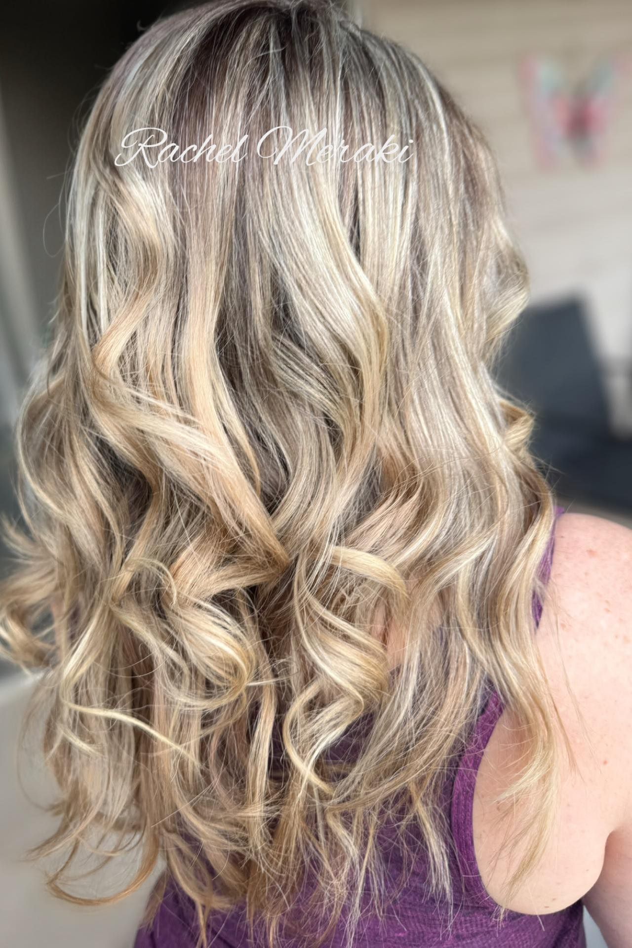Long wavy blonde hair with soft highlights, styled in loose curls against a blurred salon background