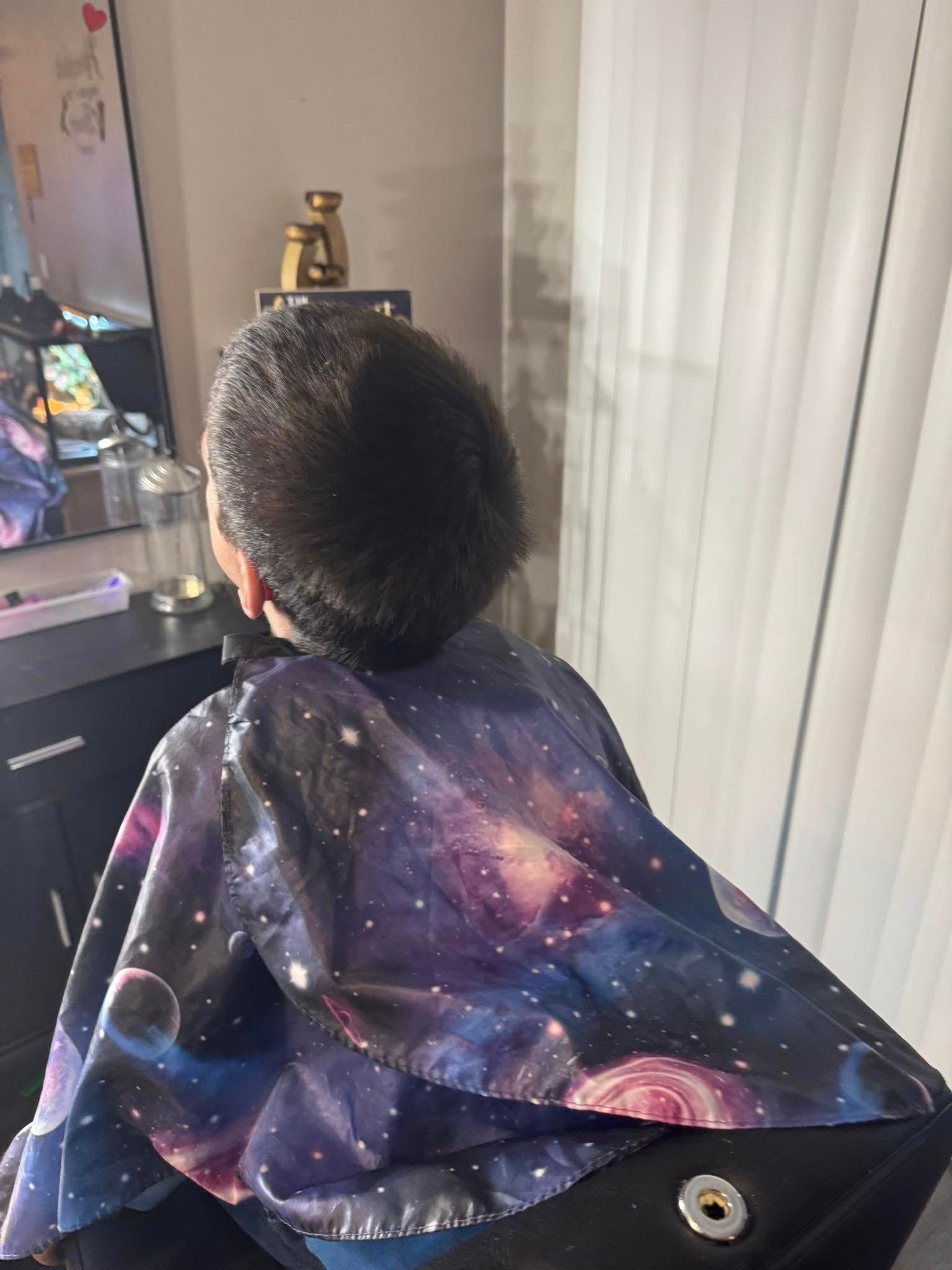 Person with curly hair wearing a galaxy-print cape, seated in a room near a mirror and blinds.