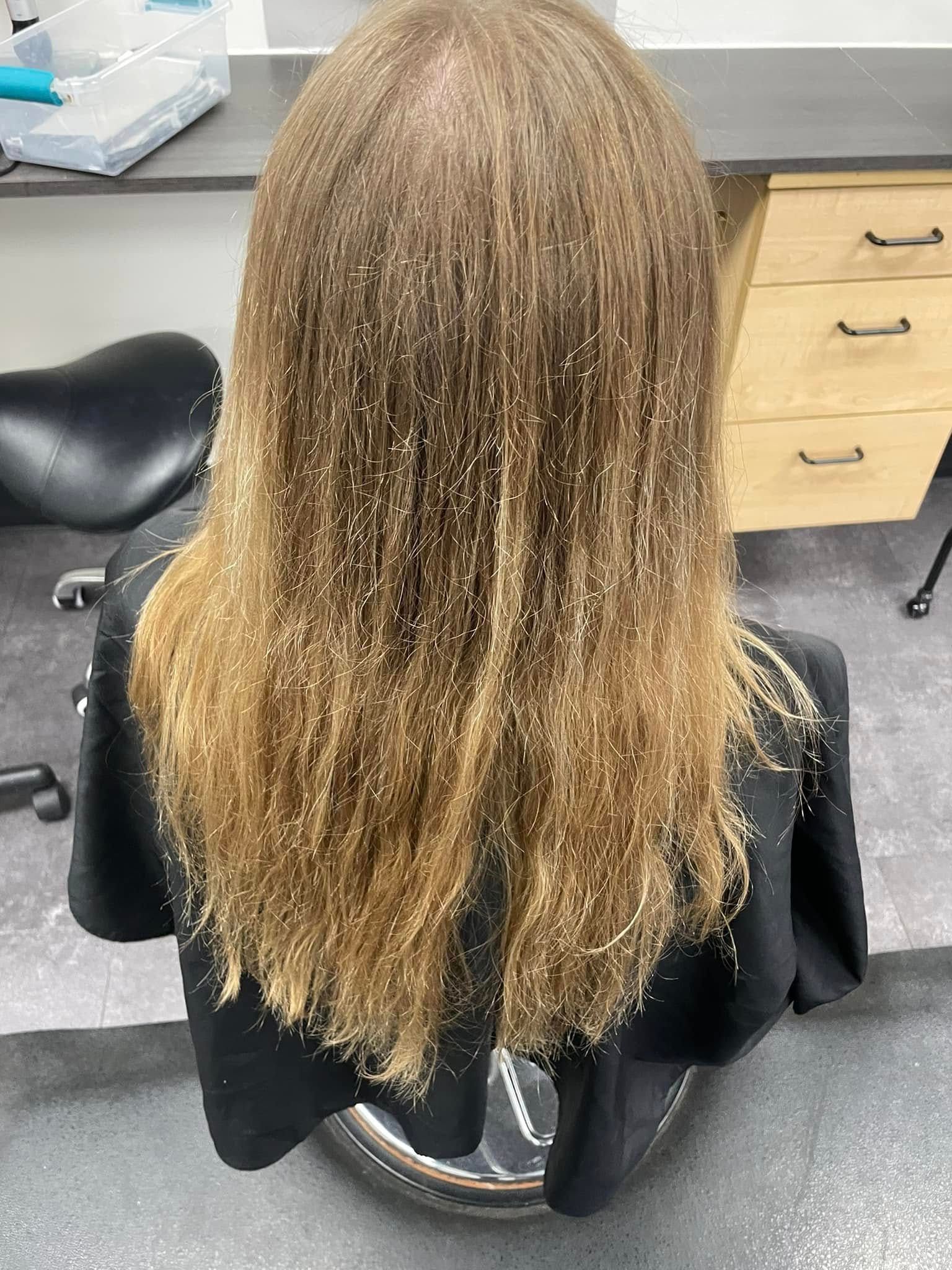 Person with long, curly light-brown hair sitting in a salon chair, viewed from behind.