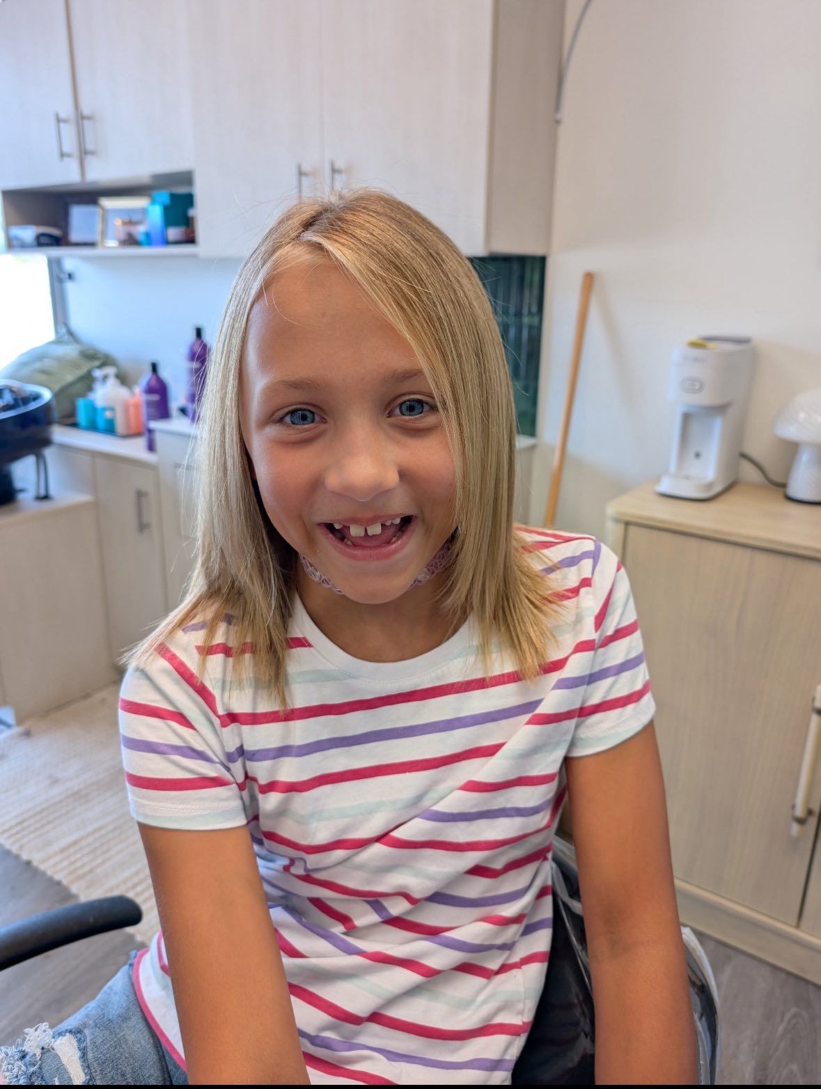 Smiling child in a striped shirt sitting indoors near a cabinet and shelves