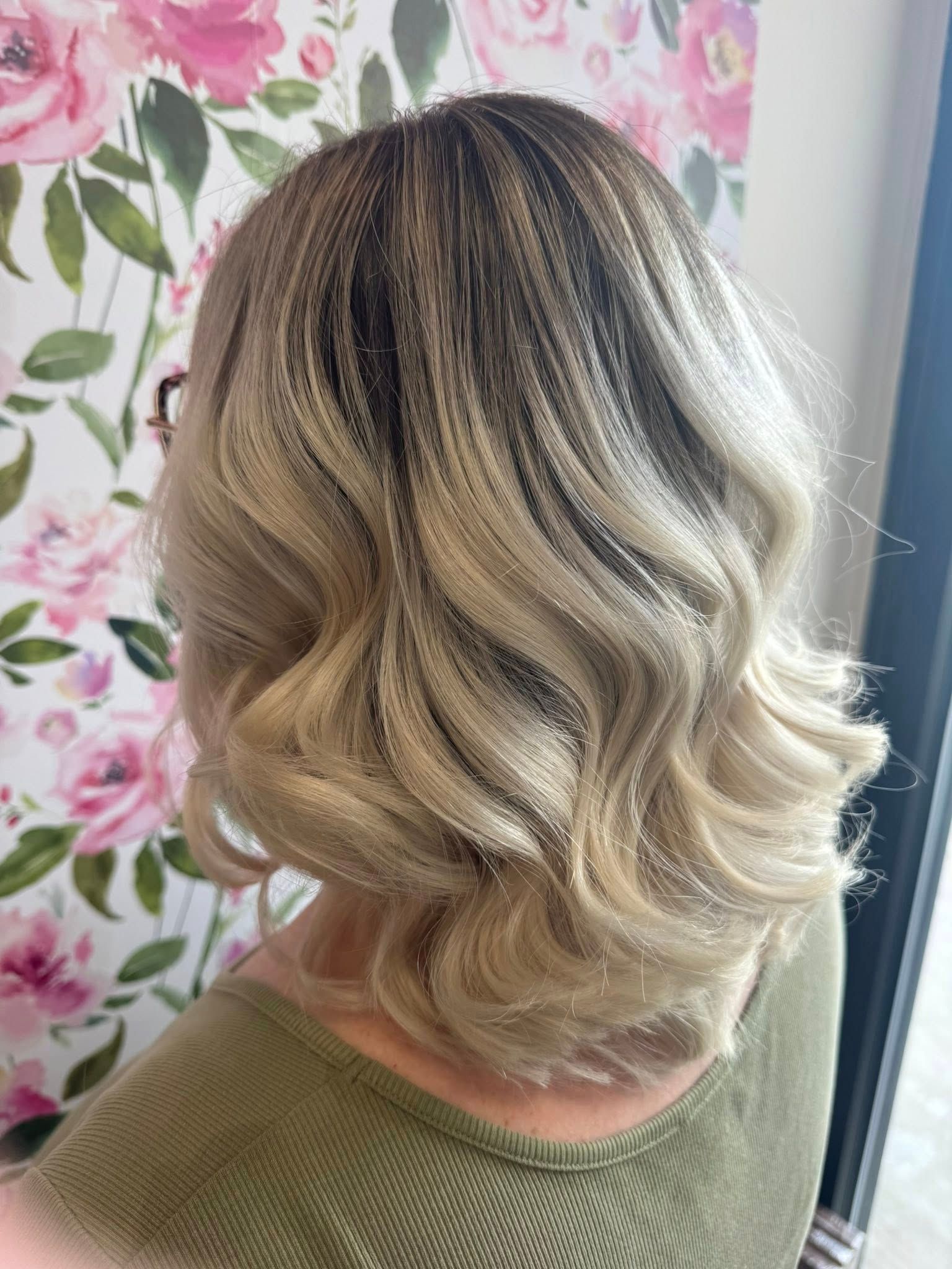 Short wavy blonde bob with dark roots, shown from the back against a floral wallpaper background