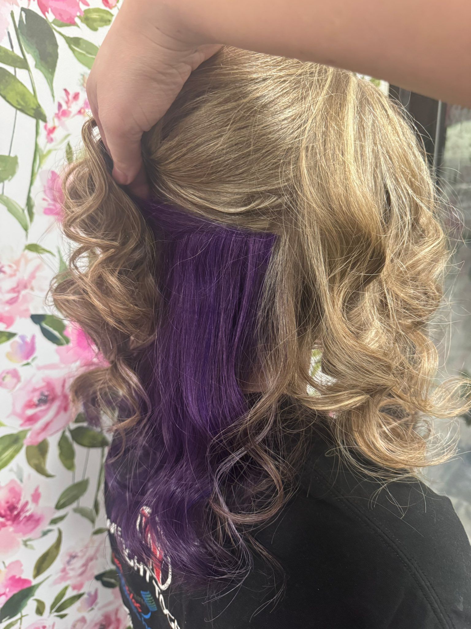 Hand holding a curly blonde wig with a purple-dyed section against a floral background