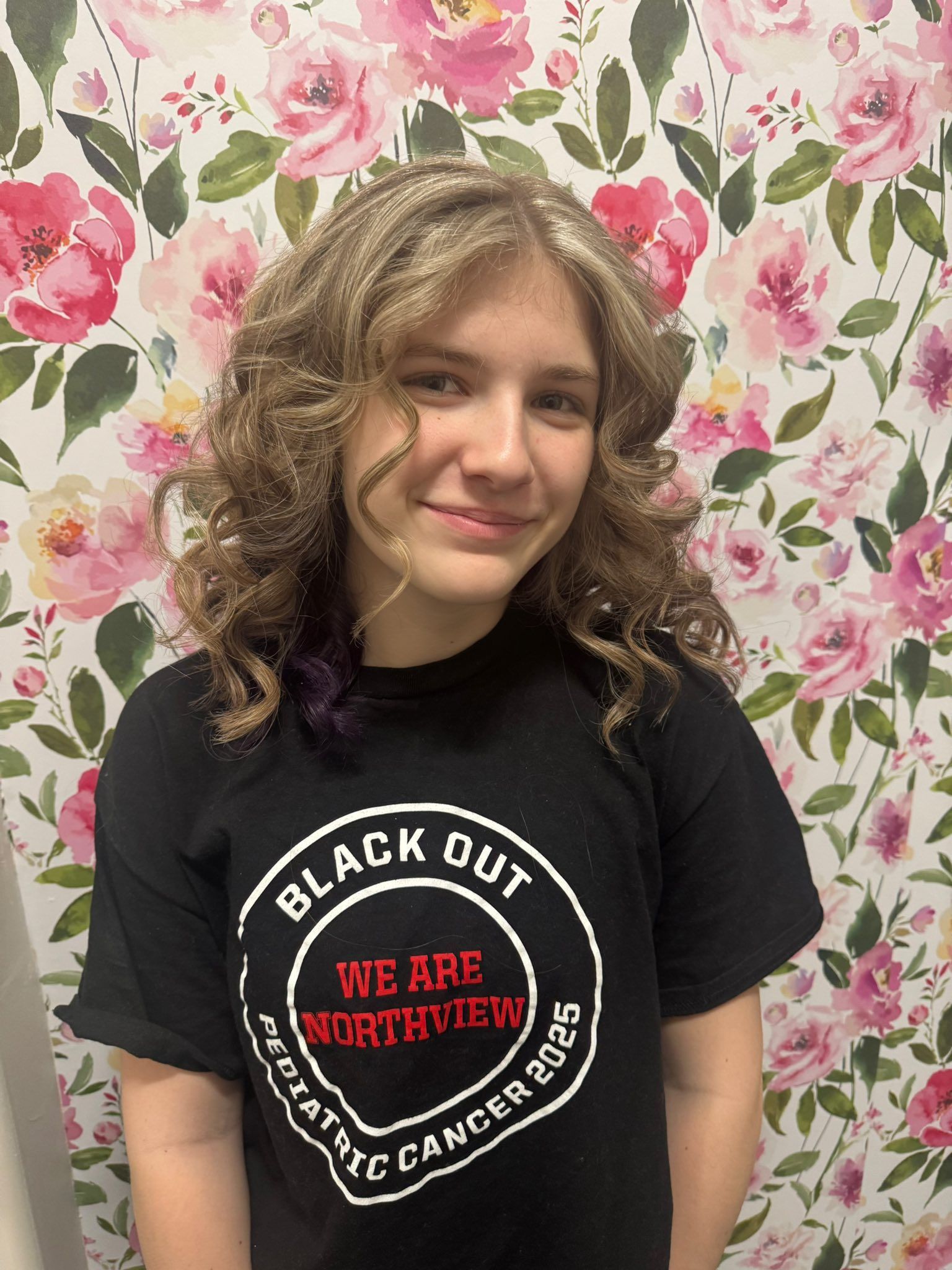 Person with curly hair wearing a black T-shirt, smiling against a pink floral wallpaper background.