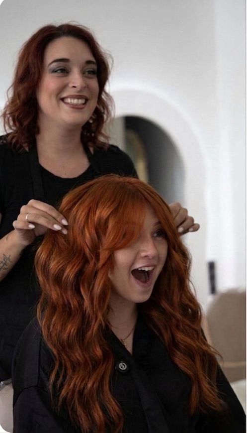 Woman styling another person’s long orange hair, both smiling in a bright indoor setting