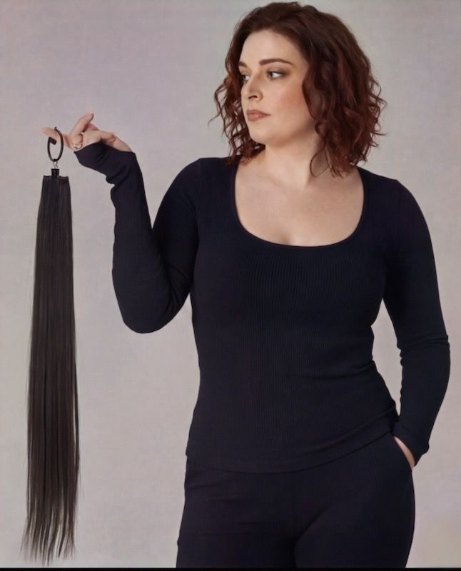 Woman in black outfit holding a long black tassel accessory against a plain gray background