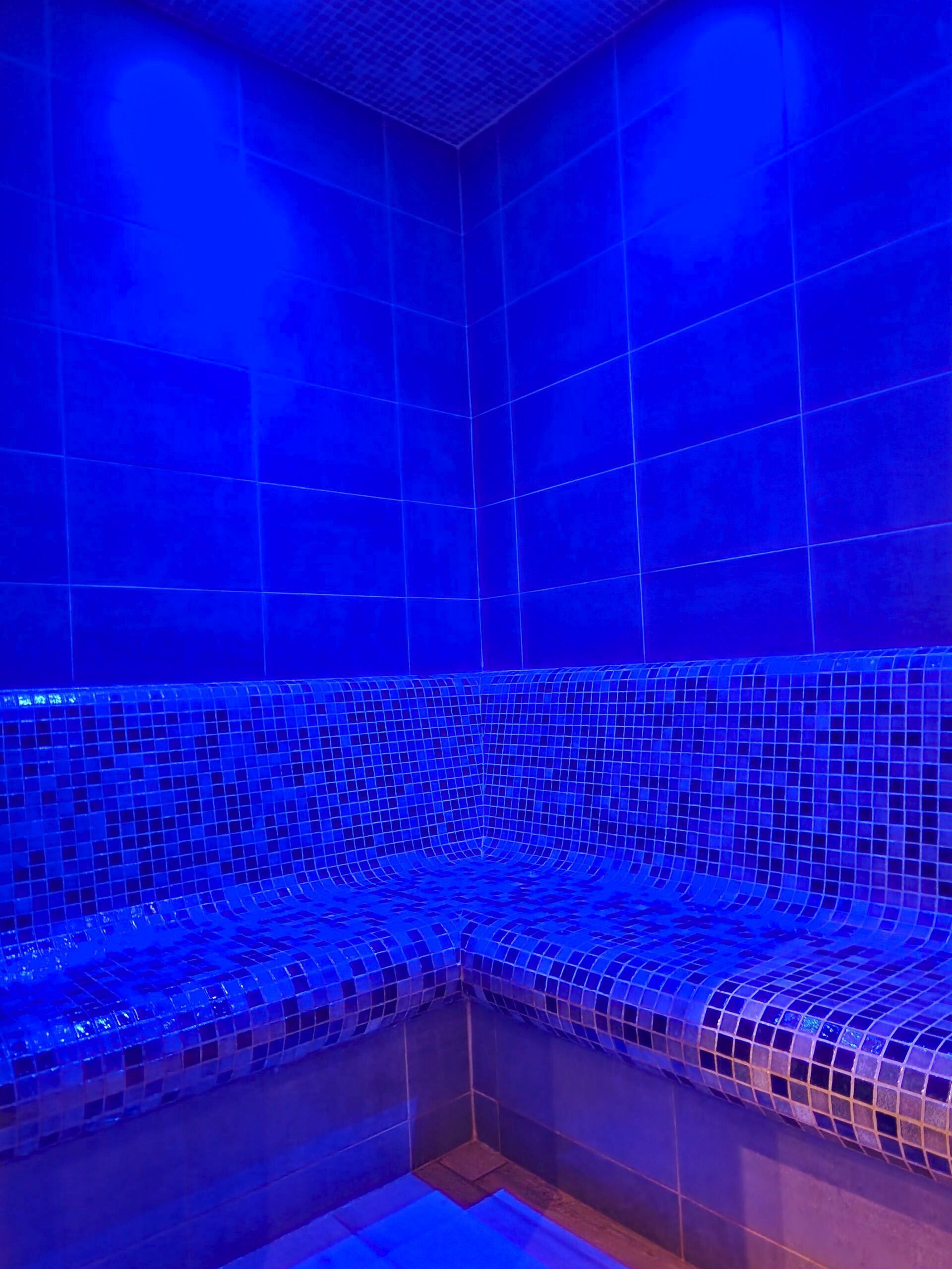 Hammam éclairé en bleu avec murs et banquettes carrelées.